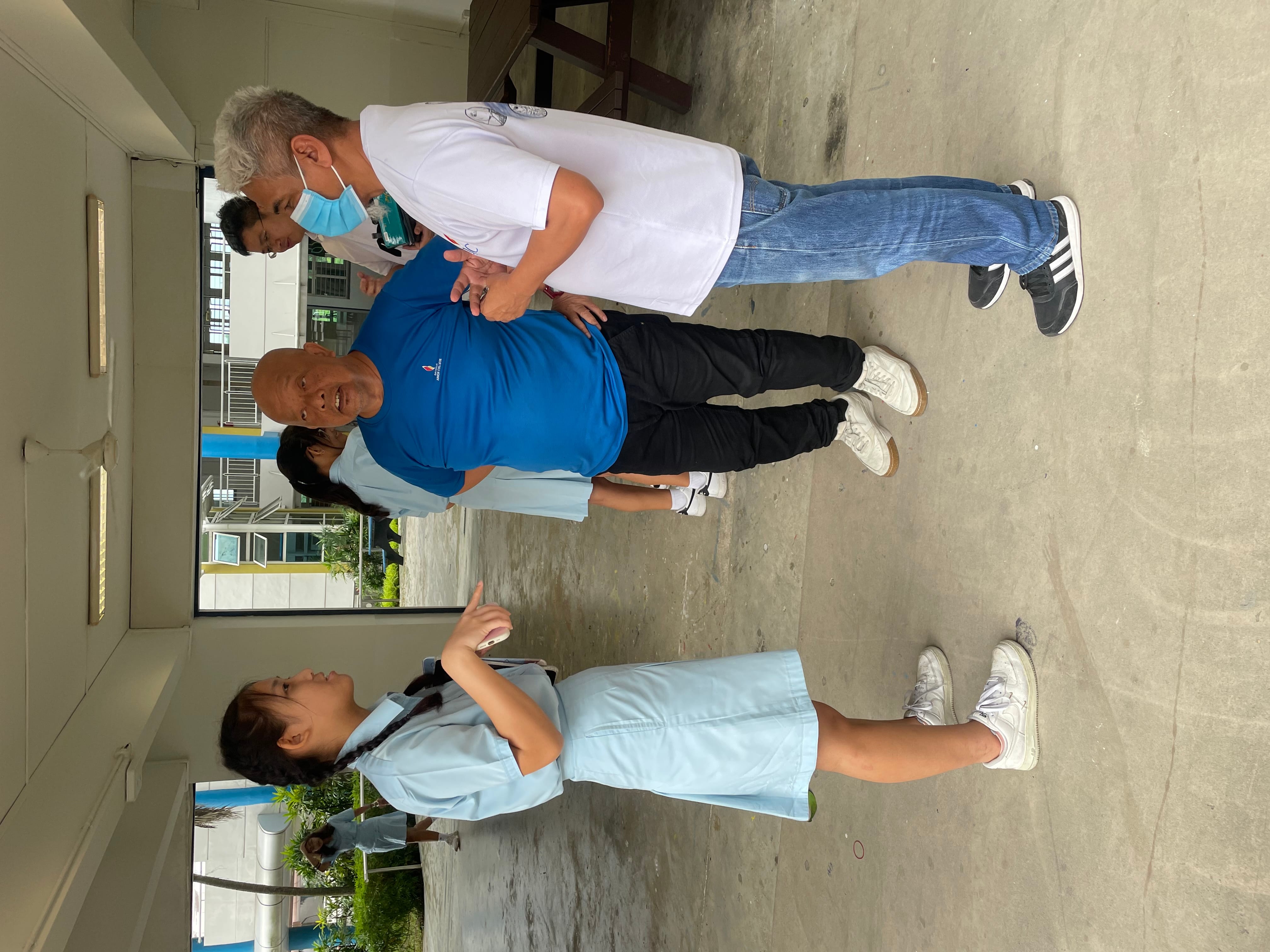 A young girl in a light blue school uniform talks with two older men in a covered outdoor area of a school.